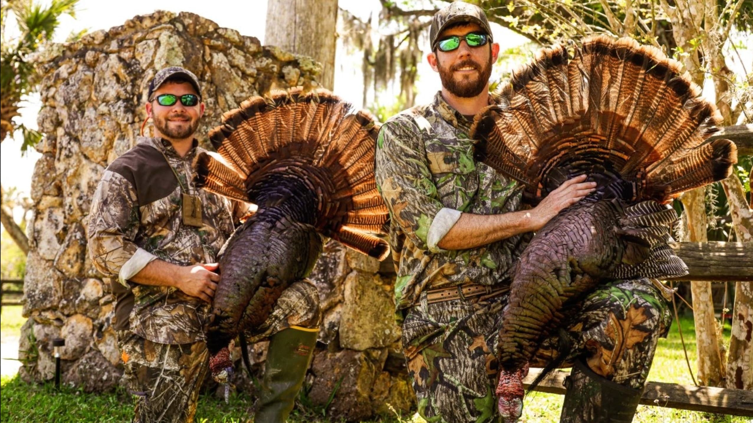 Two men in camouflage holding turkeys with fanned-out tail feathers. They stand near a stone wall, in a sunny outdoor sett...