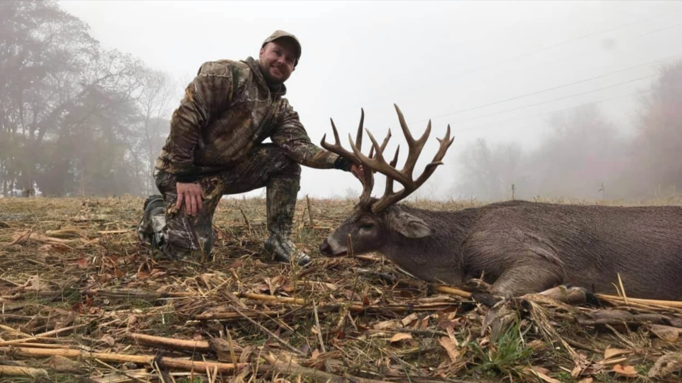 Man in camouflage kneels beside a large deer with impressive antlers, lying on the ground. Overcast day with fog in the ba...