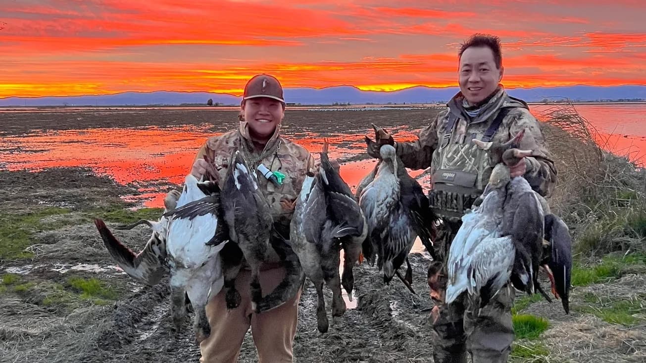 Two hunters holding harvested ducks in front of a colorful sunset over a marshy landscape.