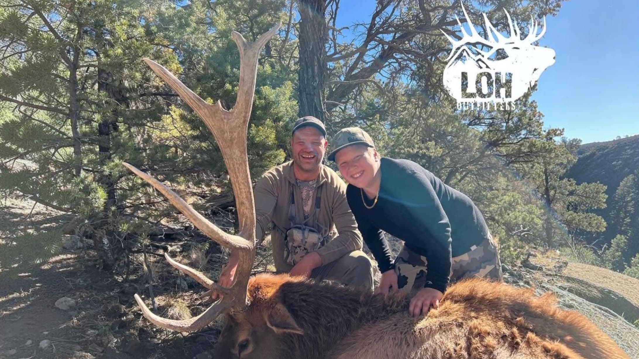 Two hunters kneeling proudly behind a harvested elk with massive antlers in a wooded mountain landscape.