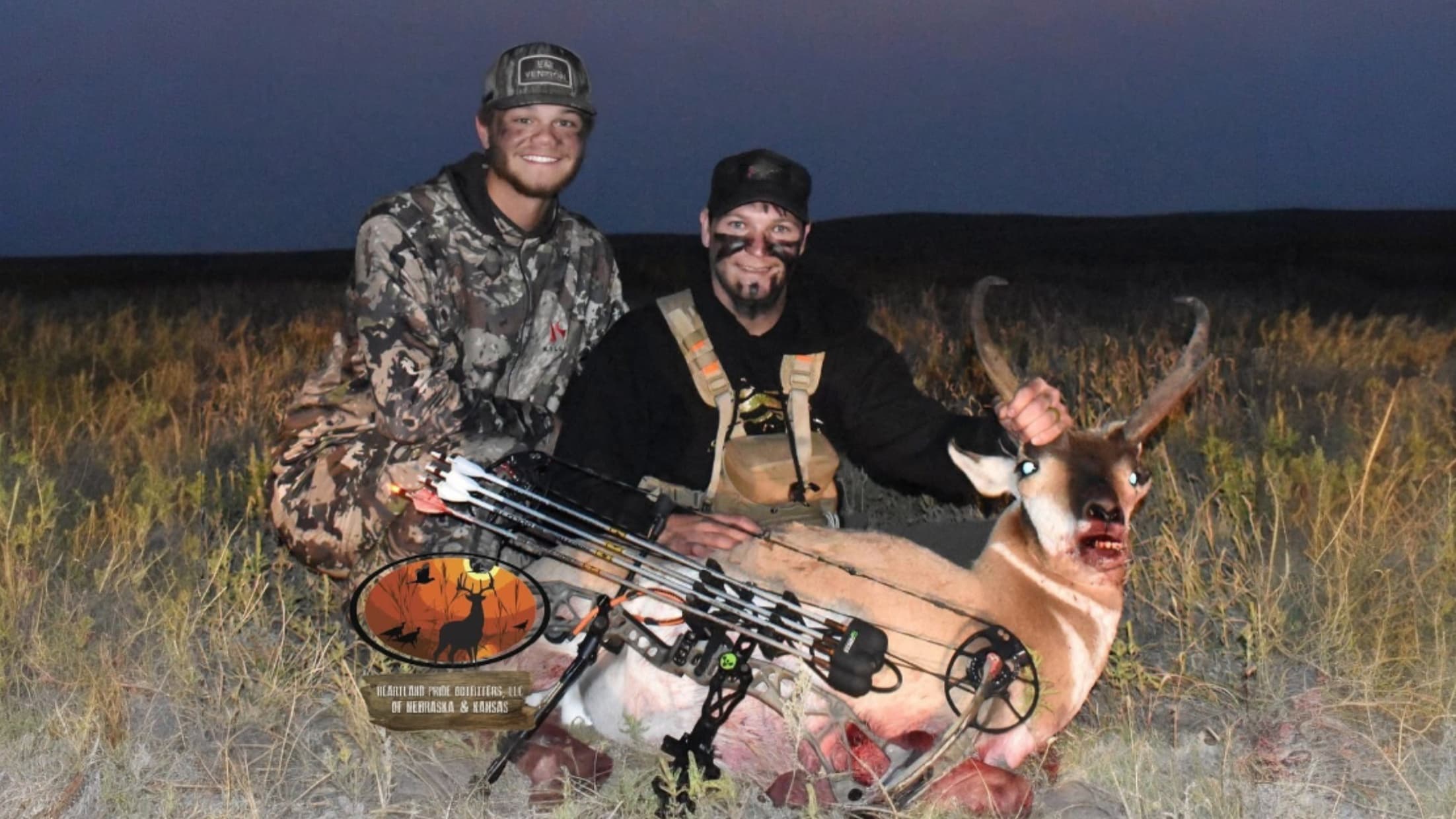 Two men pose with a hunted pronghorn antelope. One holds the antlers. Hunting bow and arrows in the foreground. They stand...