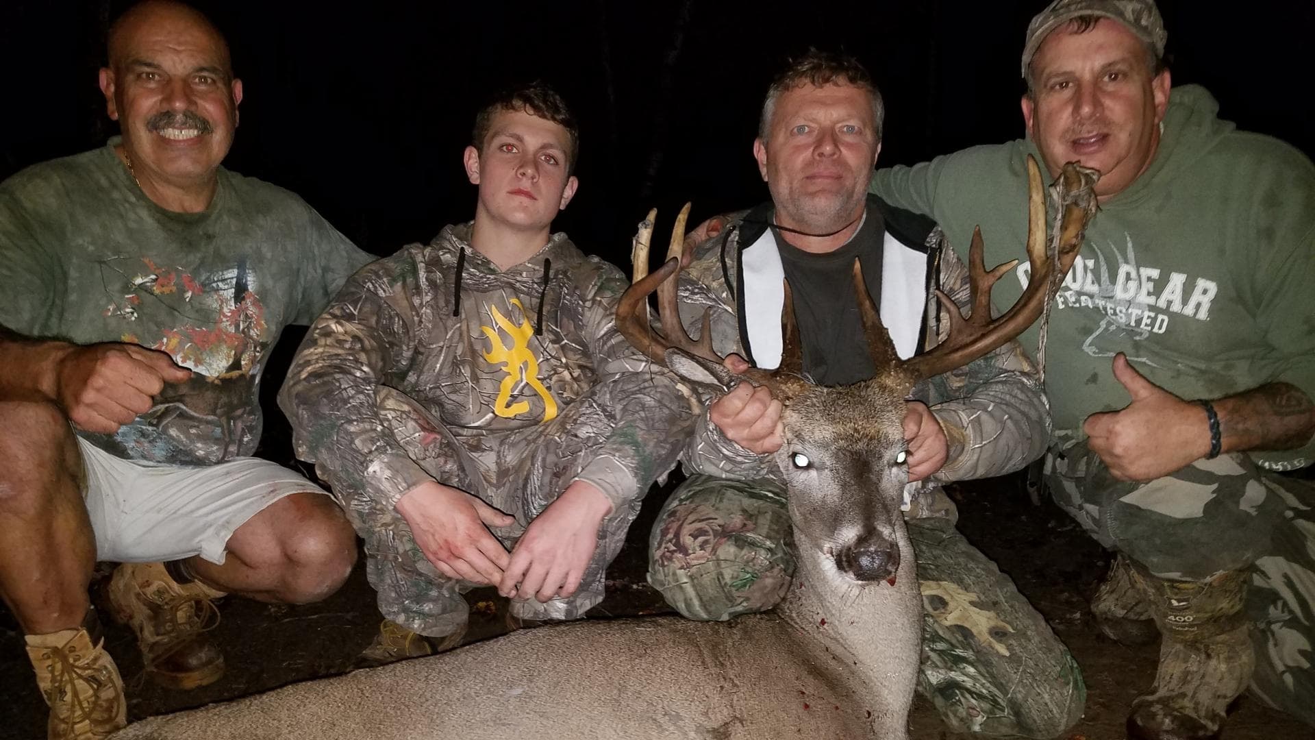 Four men pose with a large buck. The men wear hunting attire, with one holding the deer's antlers. They are crouched down ...