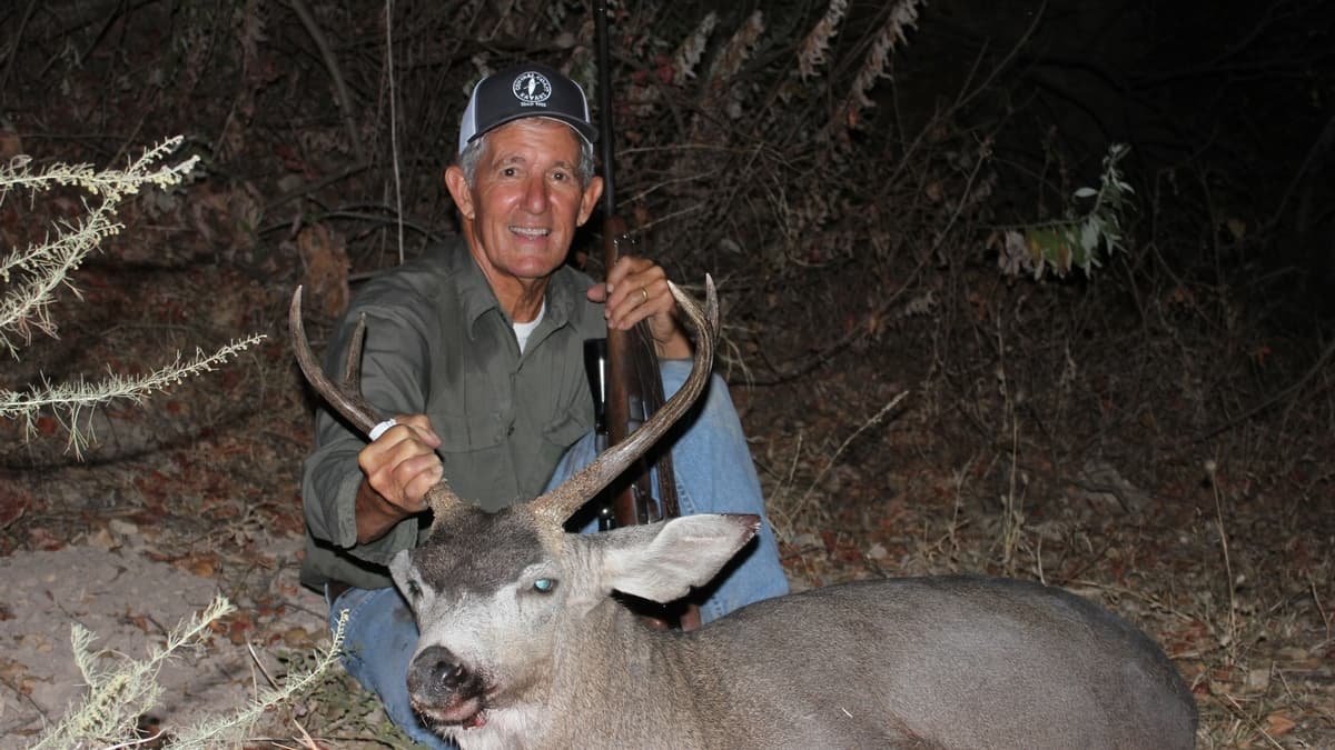 Man holding a deer's antlers, smiling at the camera. He is kneeling near the deer. A rifle rests against him. Forest backg...