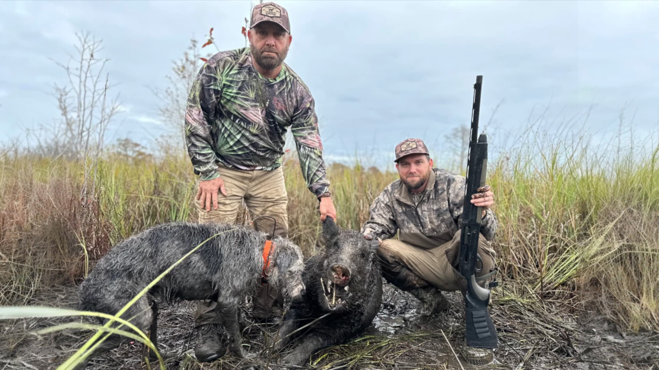 Two men in camouflage pose with a dead boar and a dog in a muddy field. One man holds a gun. Overcast sky.