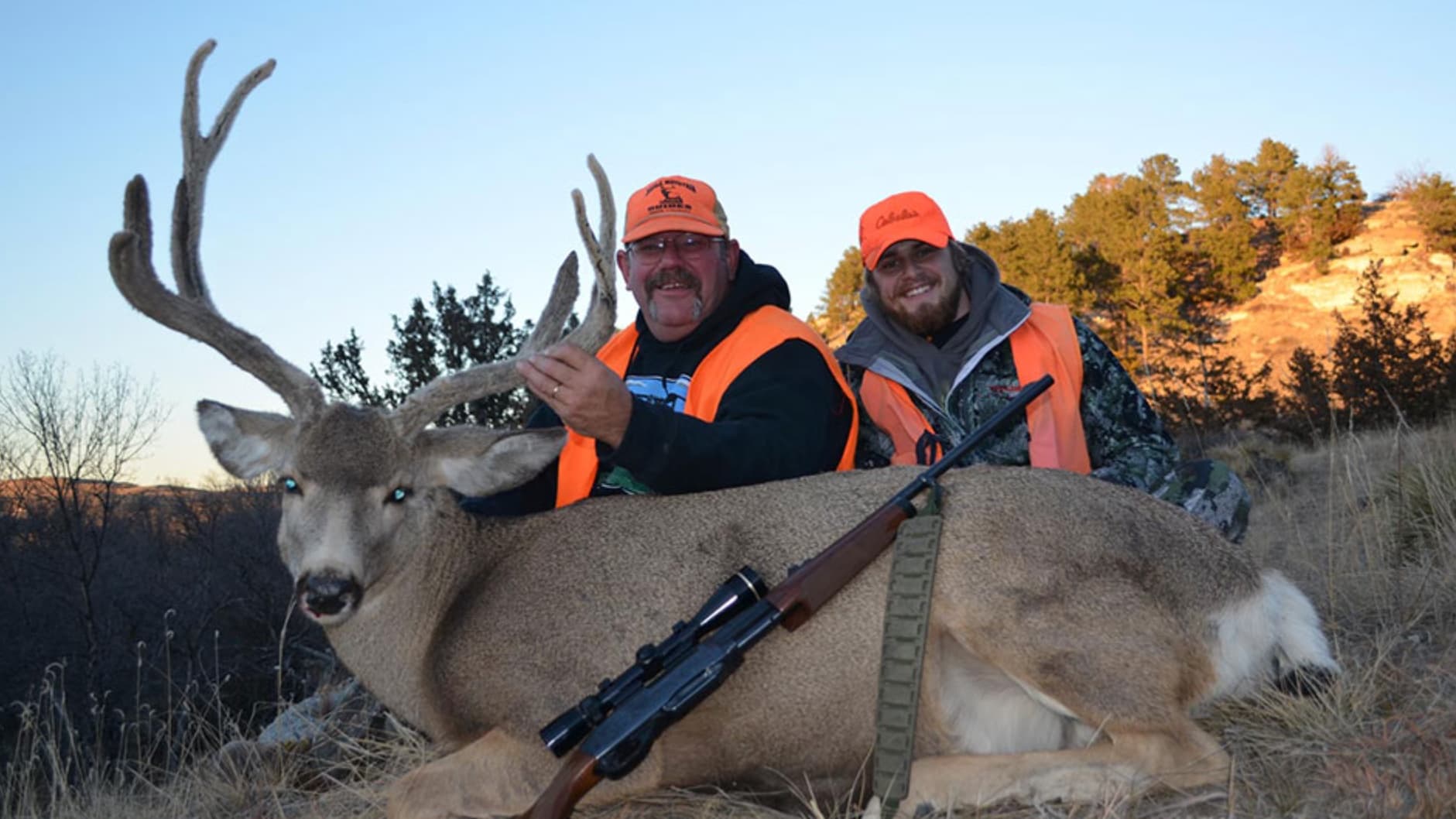 Two hunters in orange vests posing with a large buck. Rifle rests on the deer's body. Bright sky, background of trees and ...
