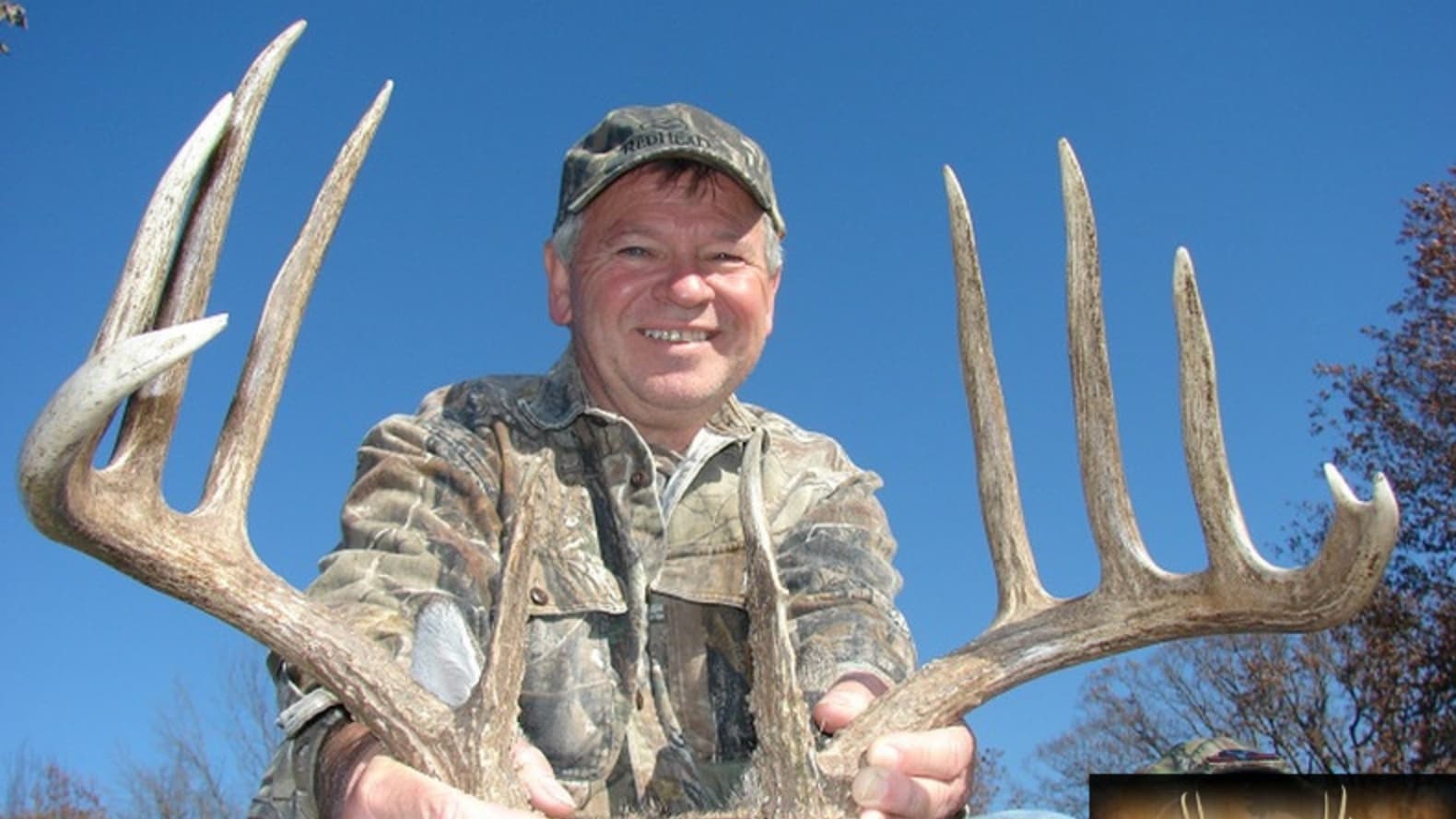 Man in camouflage holding large deer antlers against blue sky.