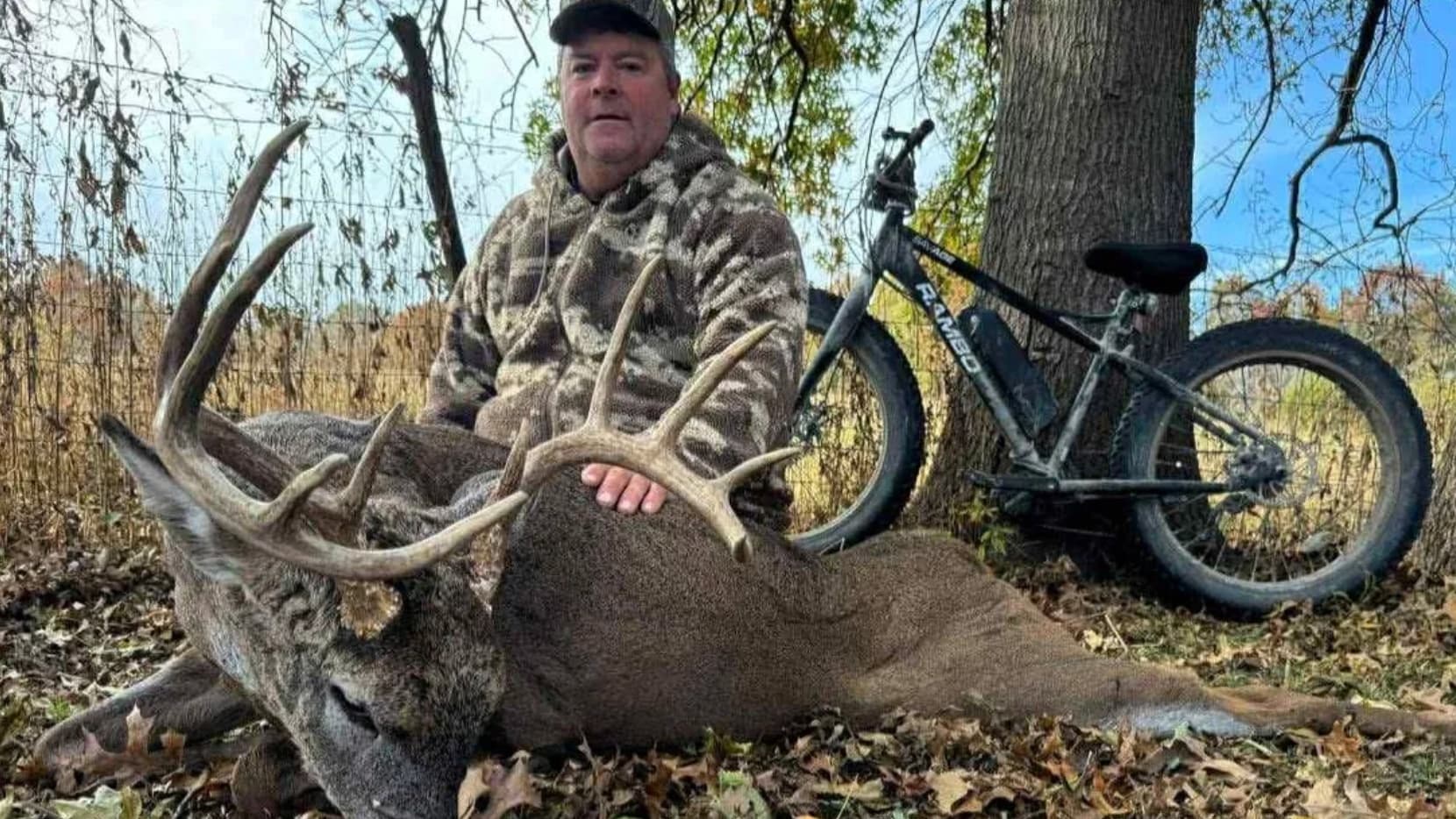 Man in camo next to large deer with antlers. A Rambo bike leans against a tree. Deer is on ground.