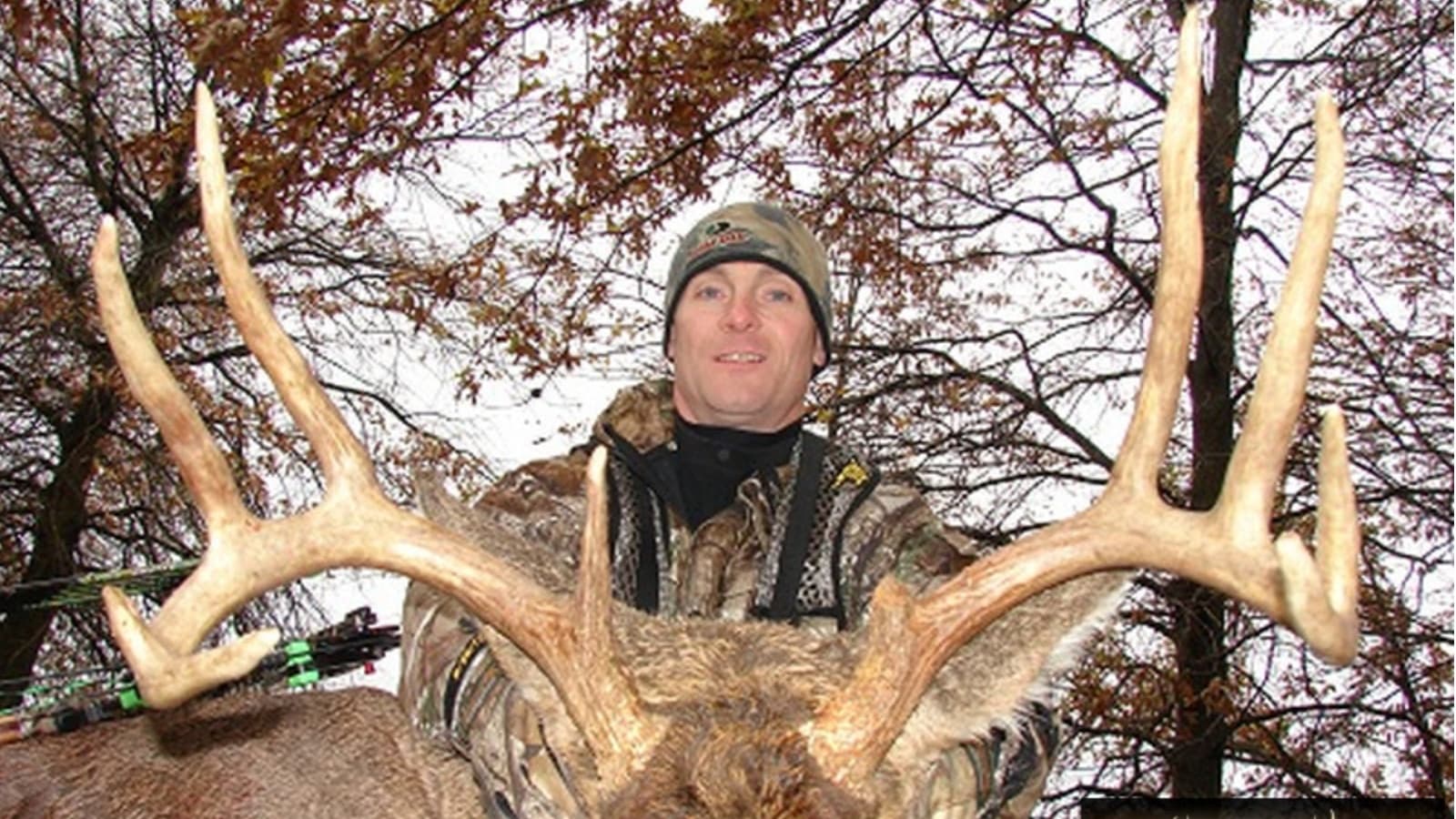 Man in camouflage holding large deer antlers, looking at the camera. Trees and foliage in the background, bow and arrow vi...