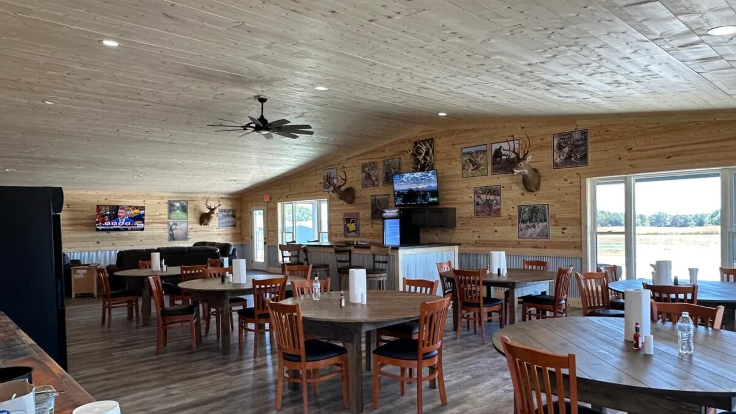 Wooden-walled interior of a restaurant with round tables, chairs, and mounted deer heads. TV on the wall, a bar, and ceili...
