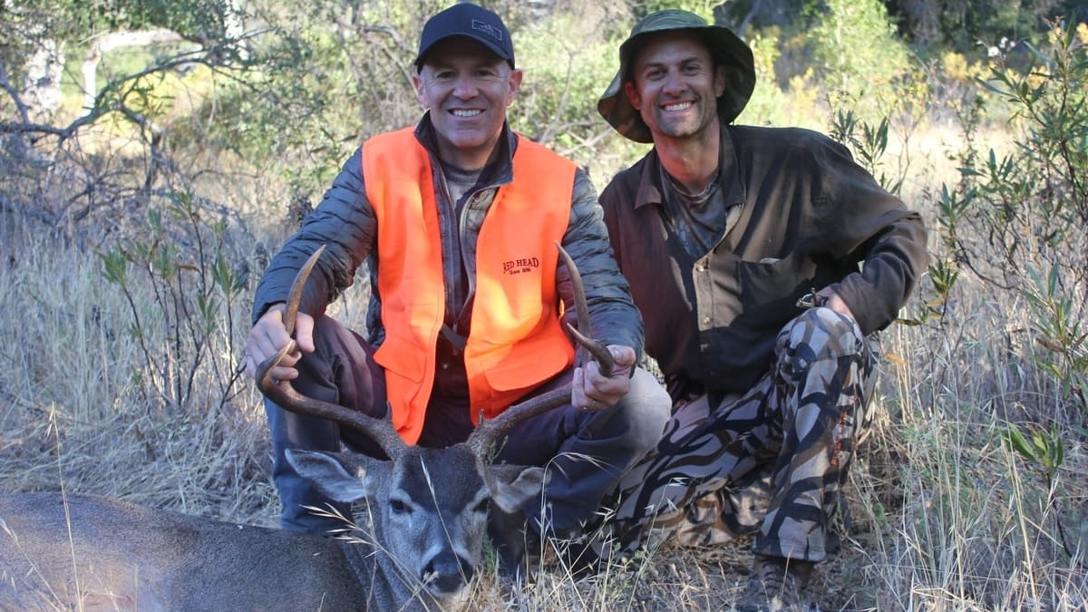 Two men pose with a buck in a field. One man wears an orange vest, holding antlers. The buck is lying down. Both men smile.