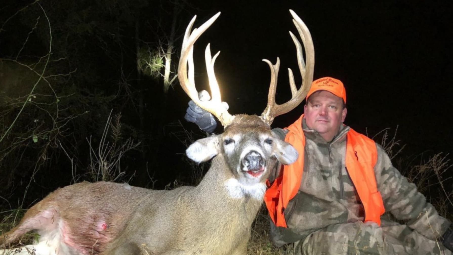 Man in hunting gear poses next to a large deer with impressive antlers. Deer's mouth shows red. Both are in a wooded area....