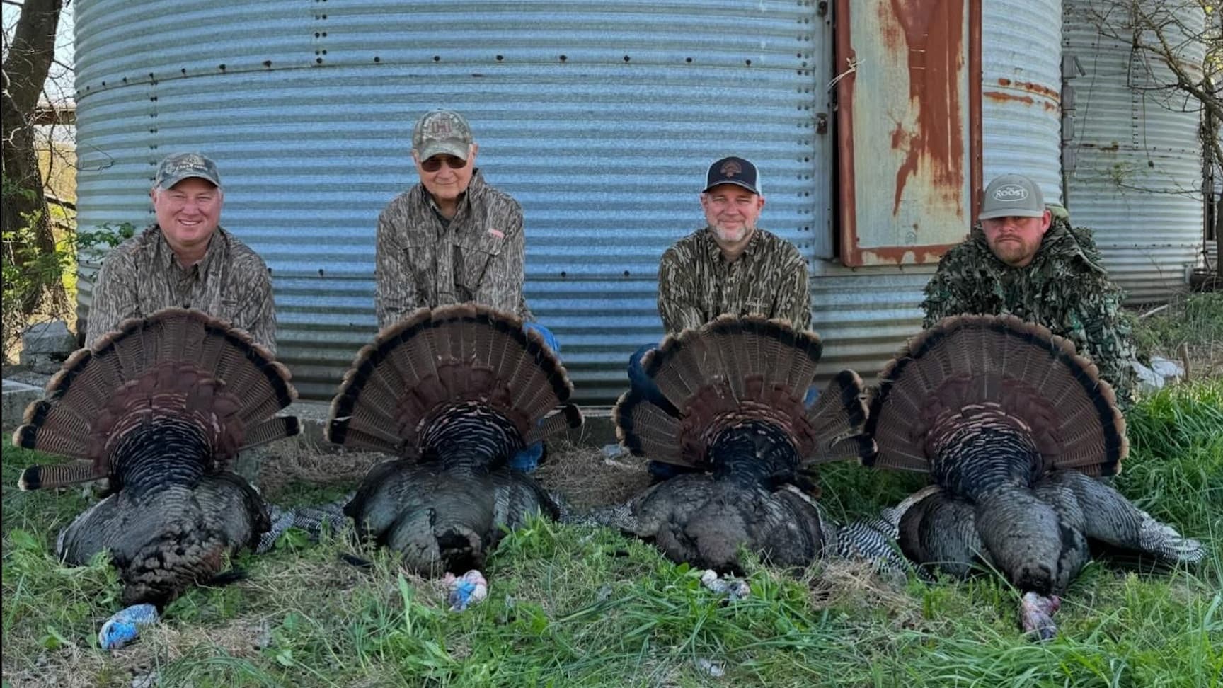 Four men in camouflage with their harvested turkeys. Each man sits behind a dead turkey with its tail feathers fanned out.