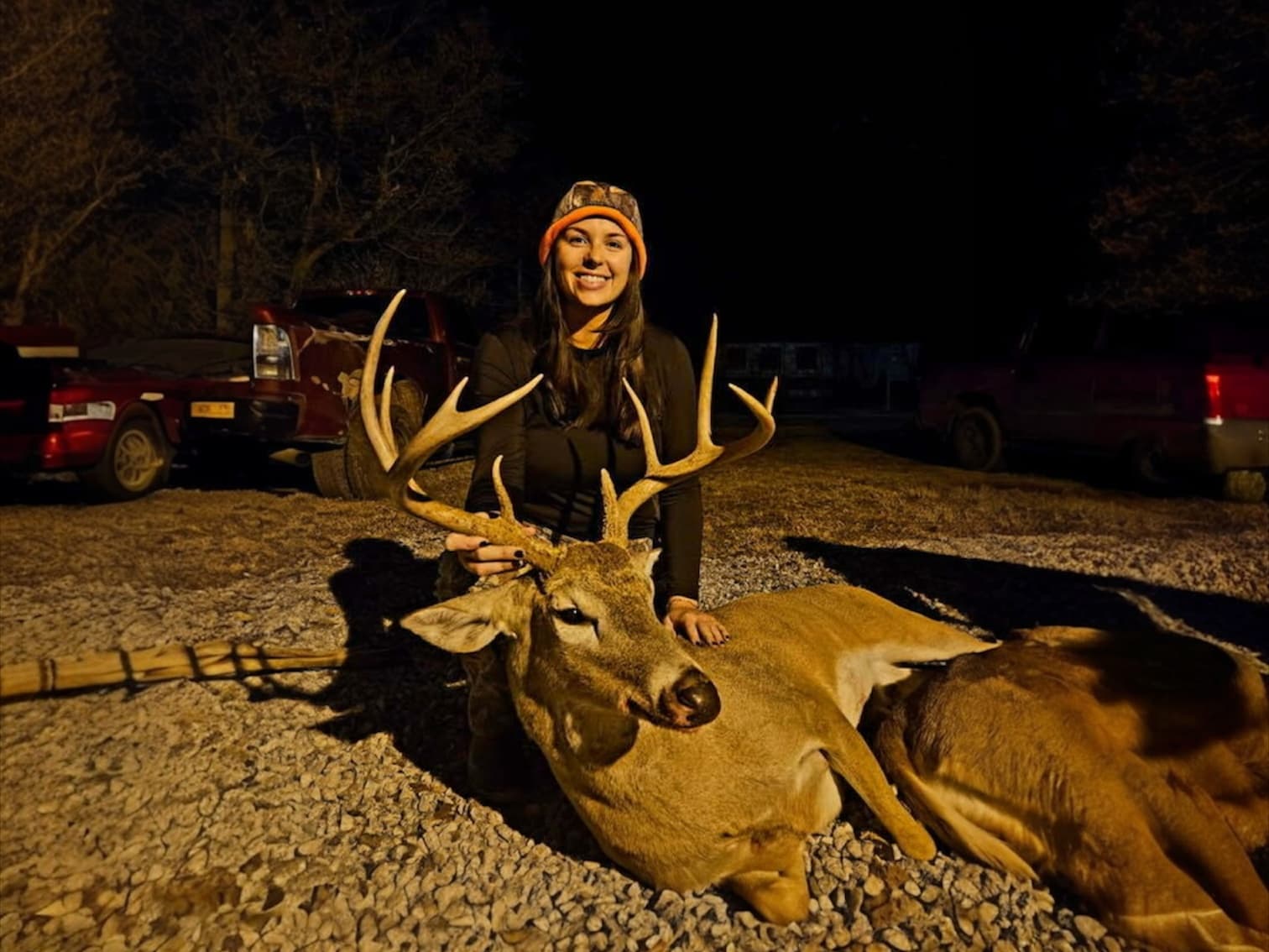 Woman with orange camo hat smiles beside a large buck, resting on gravel. Trucks are in the background. Deer has large ant...