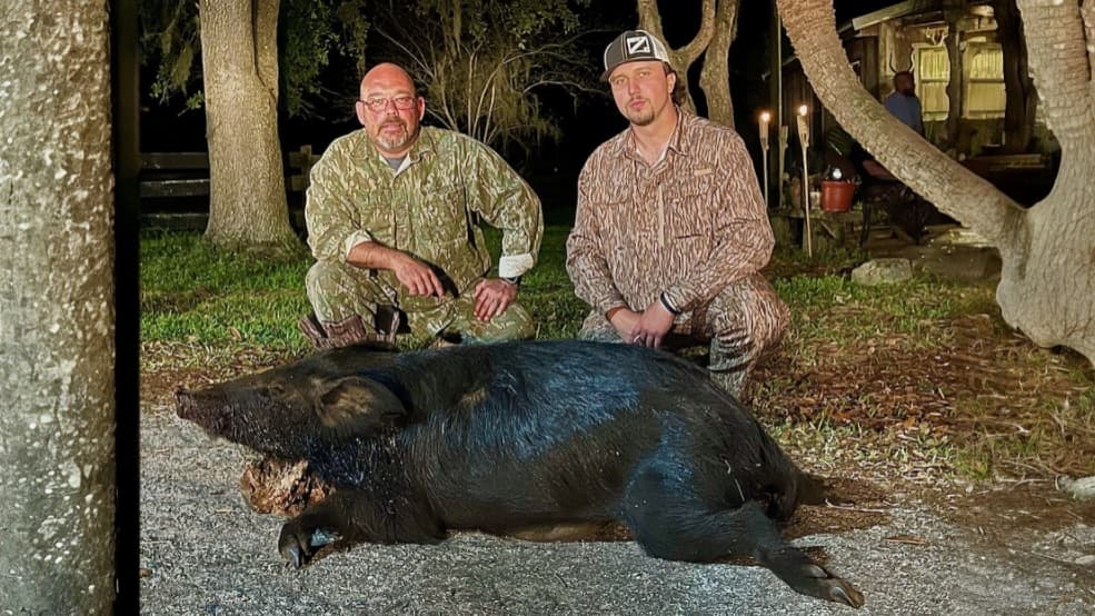 Two men in camouflage kneel beside a large, dark-colored wild boar. The boar lies on the ground. Trees and a house are in ...