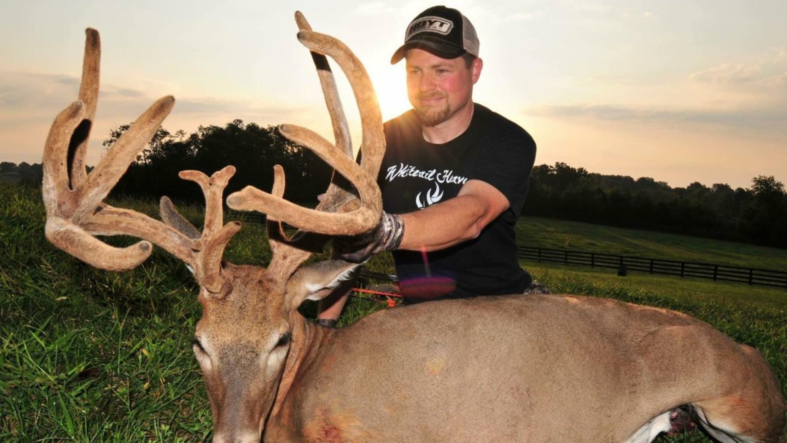 Man smiles over a large buck with impressive antlers, lying on grass. Setting sun in background. Man is wearing a cap and ...