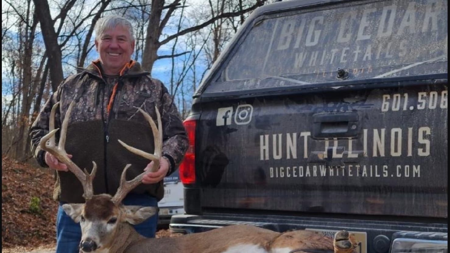 Smiling man in camouflage holds large buck near the tailgate of a truck that reads "HUNT ILLINOIS."