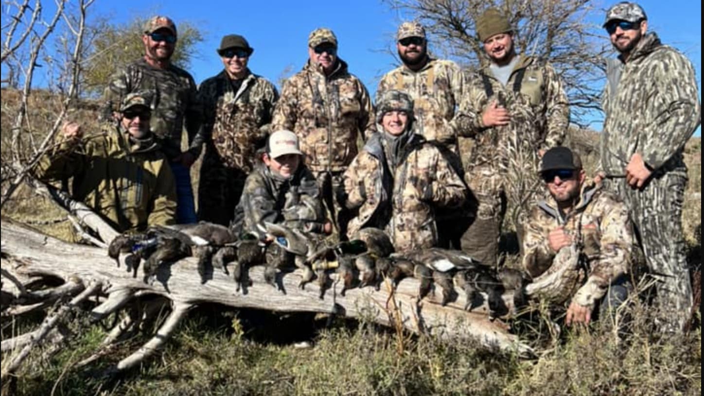 A group of men in camouflage pose with harvested ducks on a log. Men are smiling, some wearing hats and sunglasses. The la...