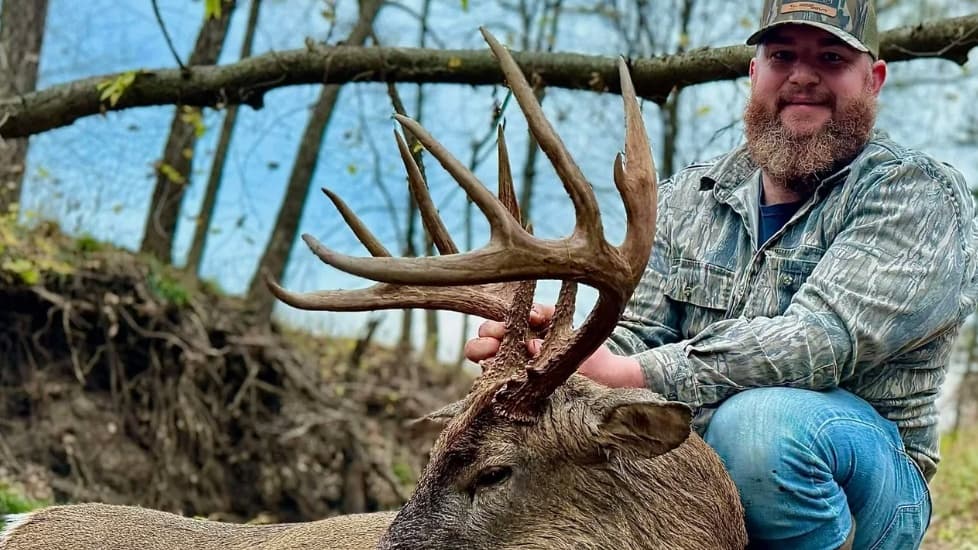 Man kneels next to a large-antlered deer, holding its antlers. He wears camo and has a beard. Trees and sky are in the bac...