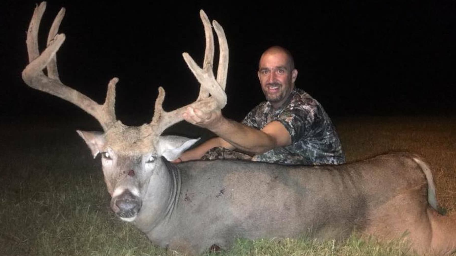 Man smiling, posing with a large buck with impressive antlers. Deer rests on ground in tall grass. Dark background.