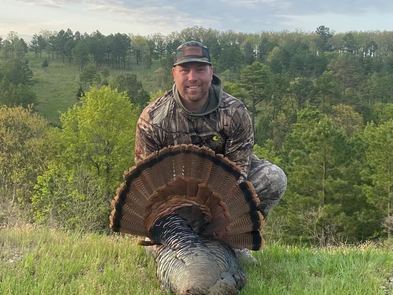 Man in camouflage kneeling, holding a wild turkey with fanned tail feathers. Forest backdrop with green trees and grassy t...