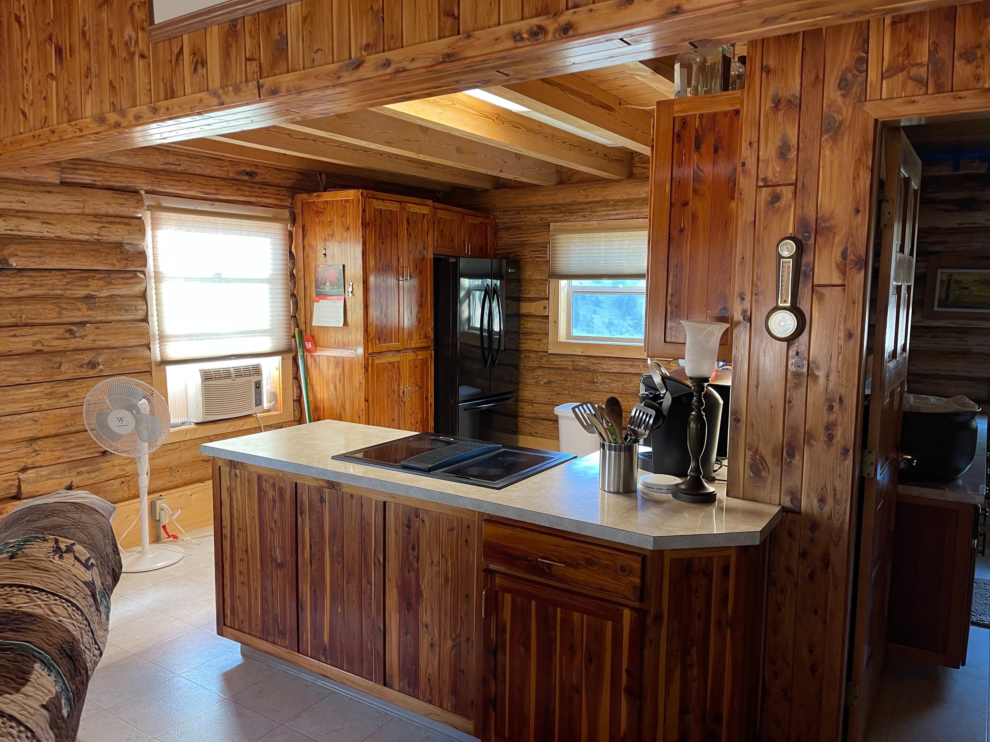 Rustic kitchen interior with wooden walls and cabinets. Center island has stovetop. Fan, window, and black refrigerator al...