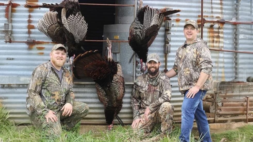 Four men pose with harvested turkeys in front of a metal building. Turkeys are hanging from hooks. Three men are in camo....