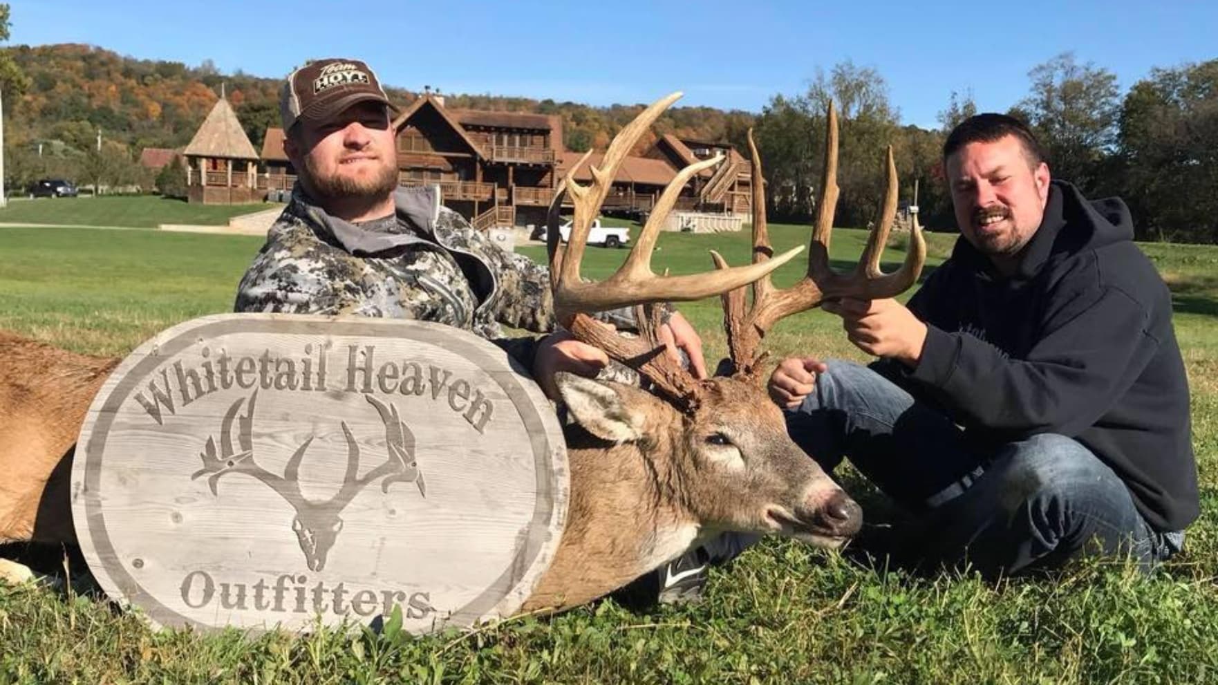 Two men with a large buck deer, antlers prominent. They're holding a wooden sign that says "Whitetail Heaven Outfitters." ...