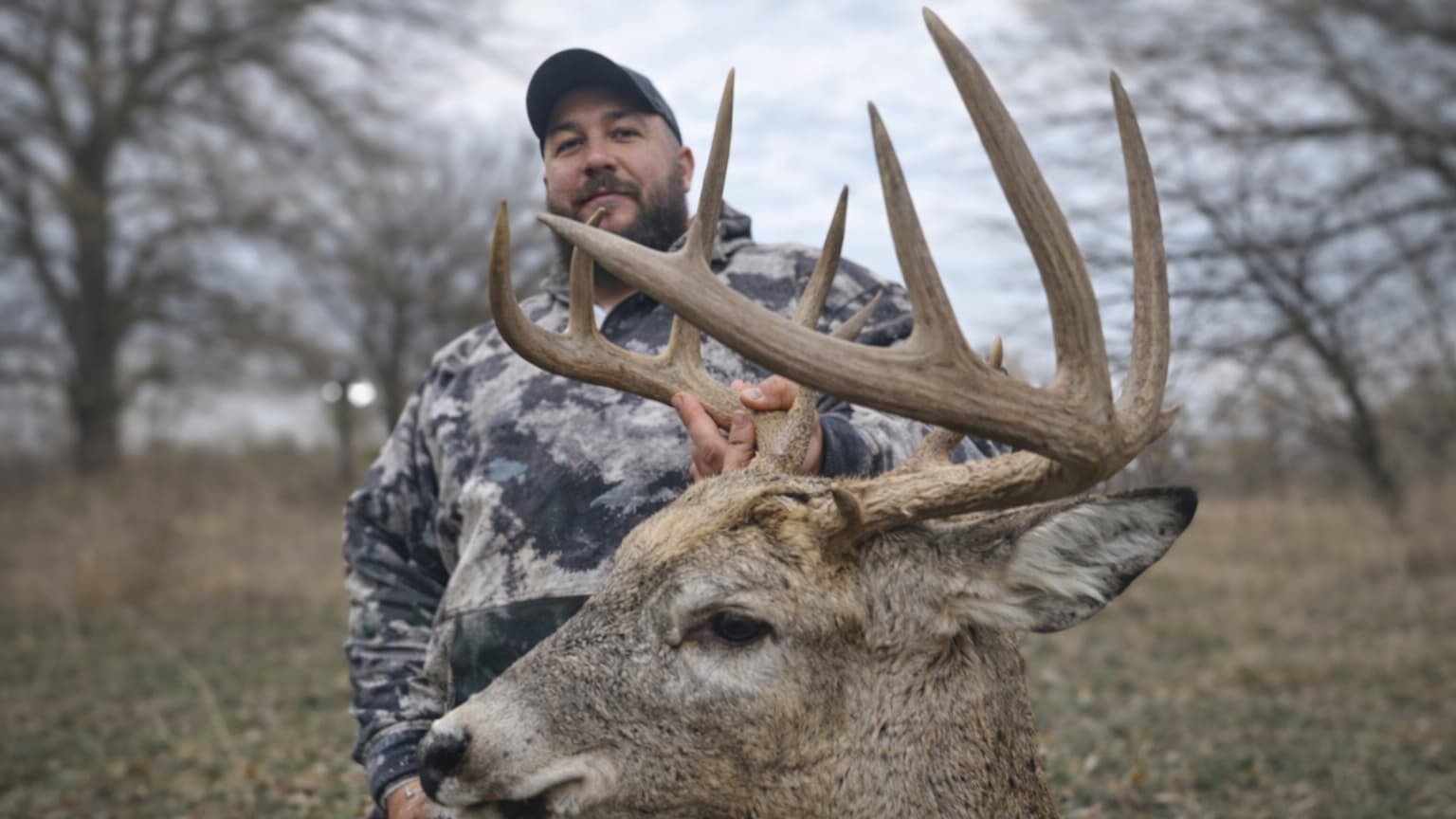 Man in camo holding the antlers of a large buck. Buck's head in the foreground, man in the background. Cloudy sky, trees.