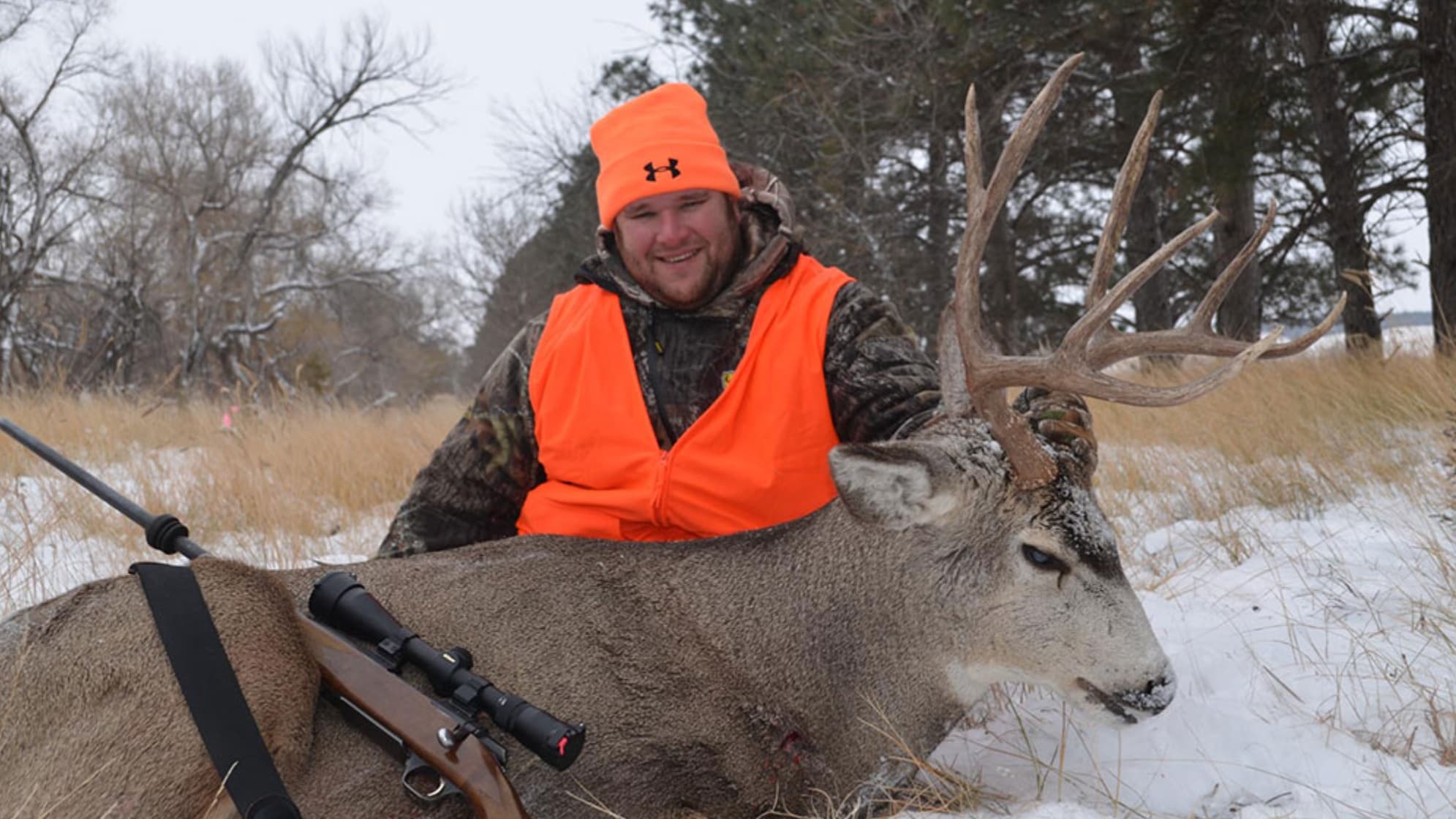 Man in orange hunting vest and hat smiles next to a large deer with impressive antlers, lying in snow. Rifle visible.