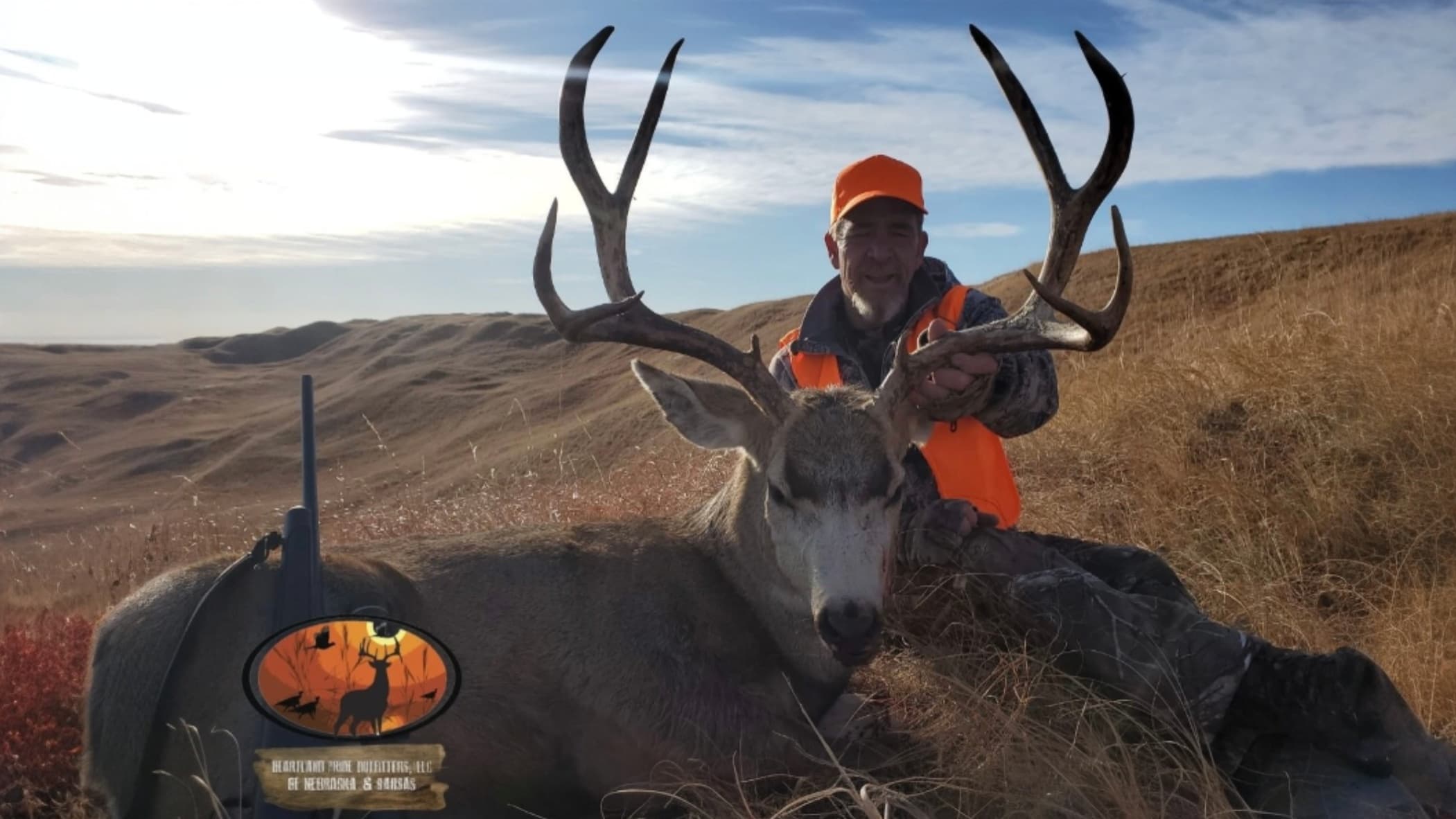 Hunter in orange vest smiles, holding a large buck's antlers. The deer lies in tall grass, rifle nearby. Landscape of hill...