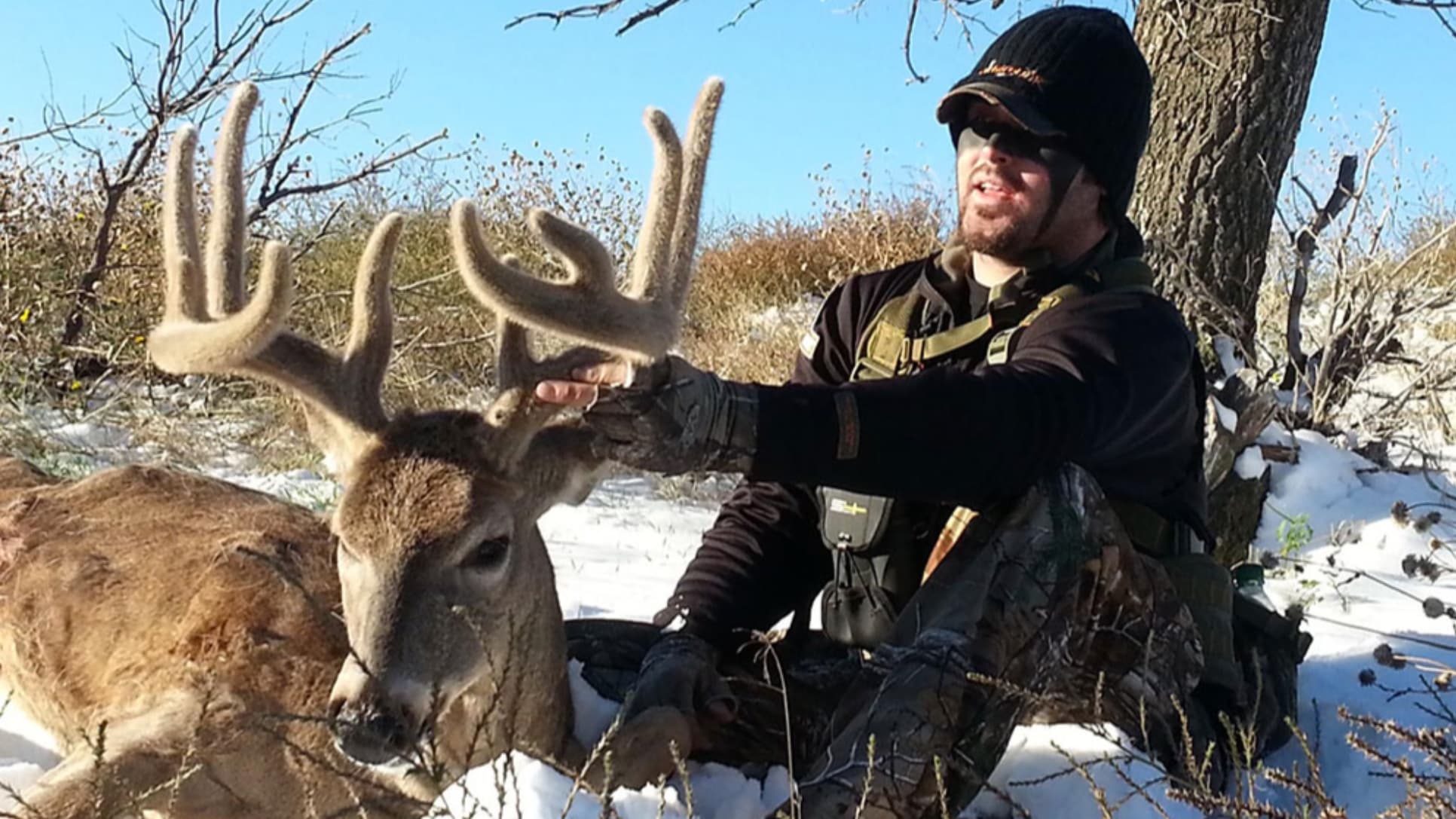 Hunter sitting with a large buck. Hunter wearing camo and a beanie, petting the deer's large antlers. Snow on the ground, ...