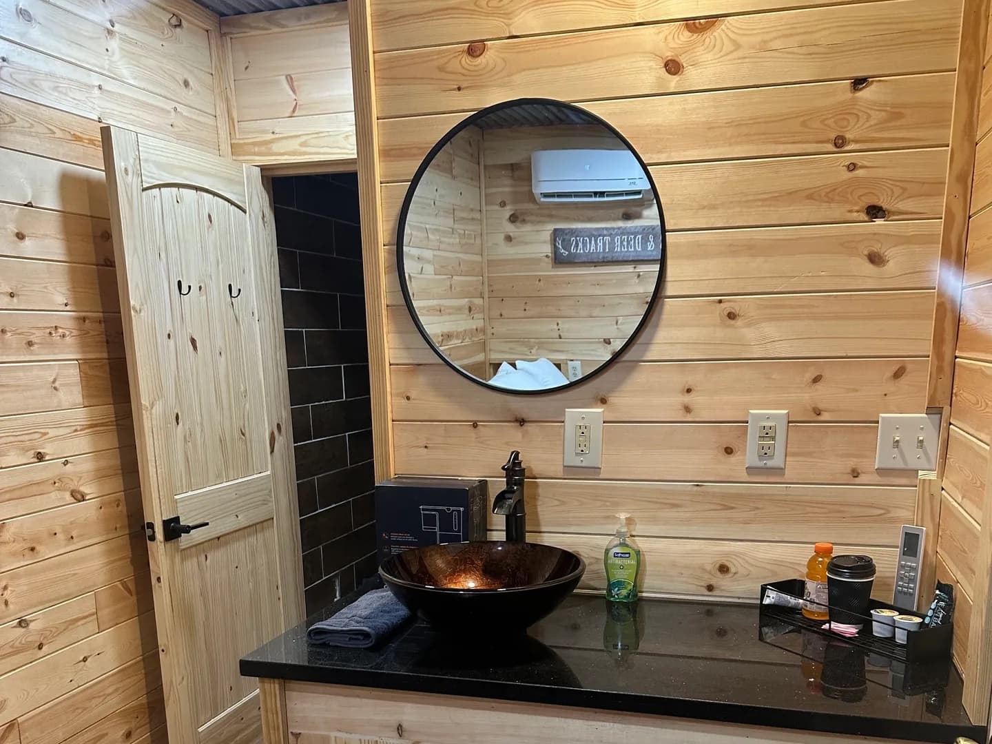 Rustic bathroom with wood-paneled walls, a black granite countertop with a dark bowl sink, and a round mirror. Open doorwa...
