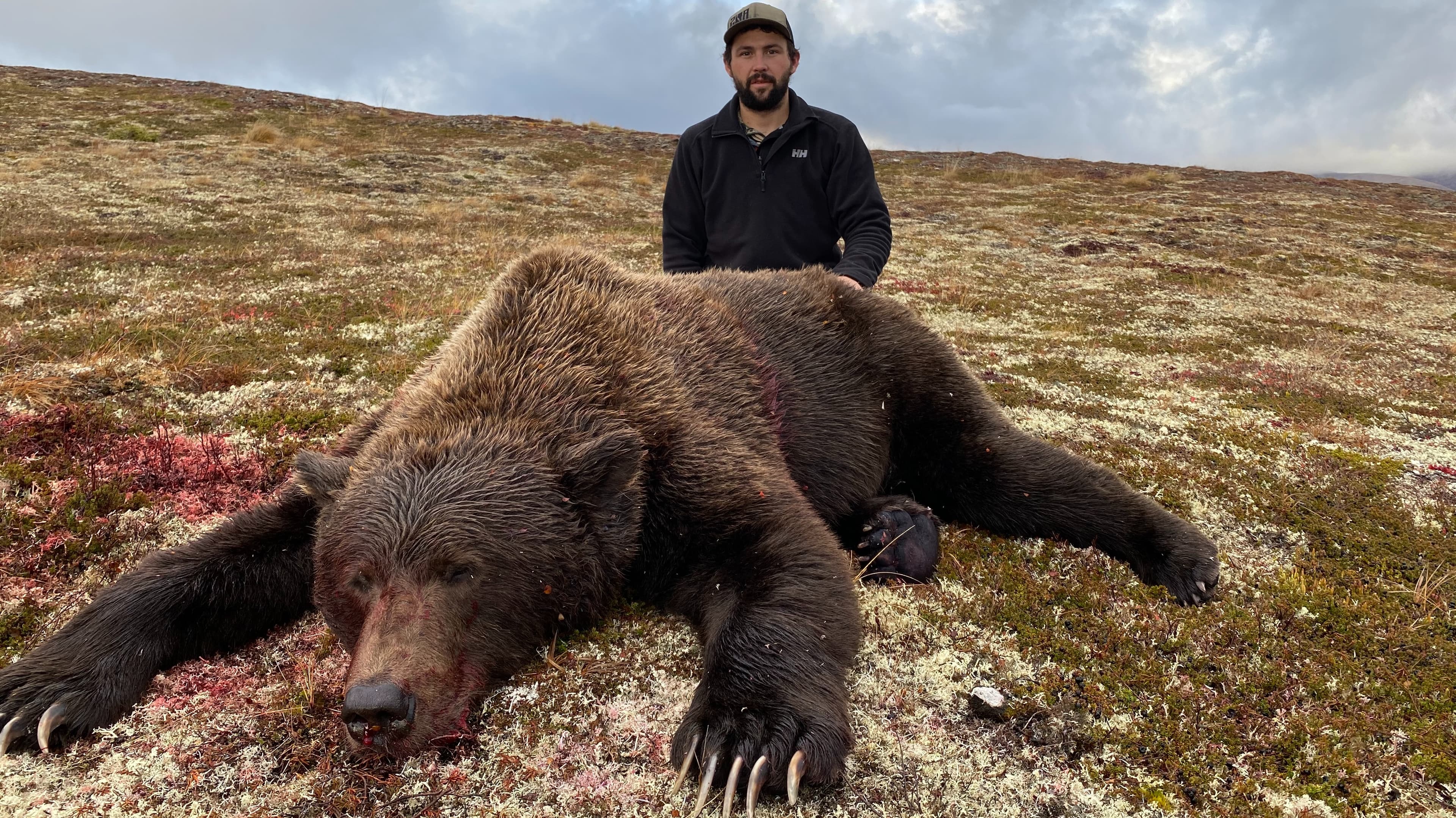 Man standing behind a large, dead grizzly bear lying on a mossy hillside. The bear has dark fur and blood on its face. Ove...