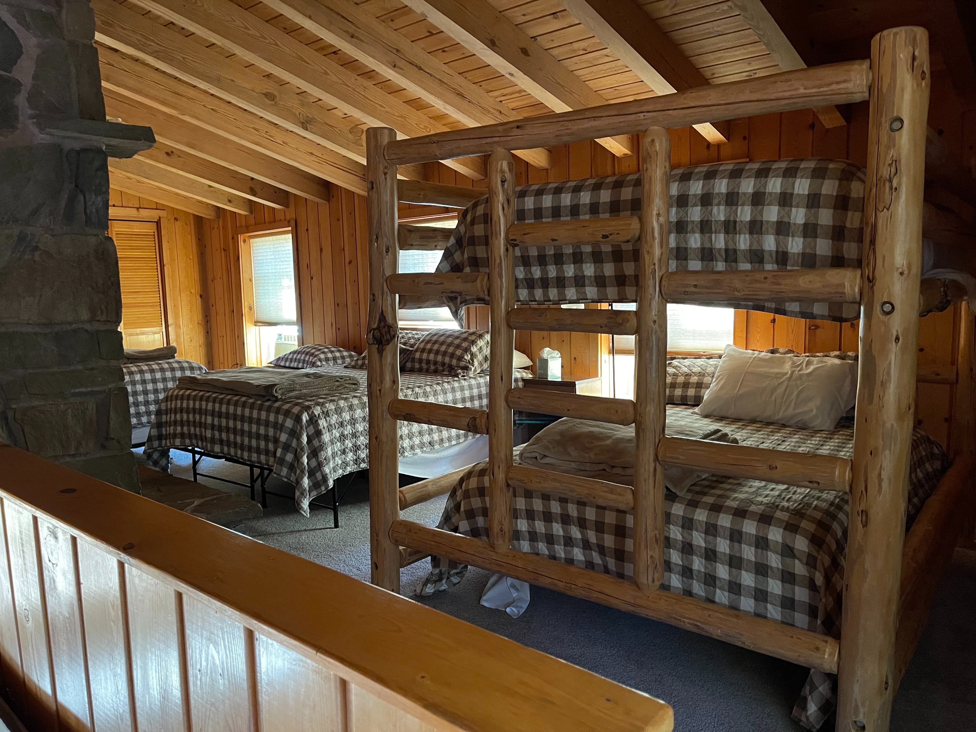 A rustic bedroom with wooden walls and ceiling. Bunk beds made of logs are the main focus, covered in brown and white pla...