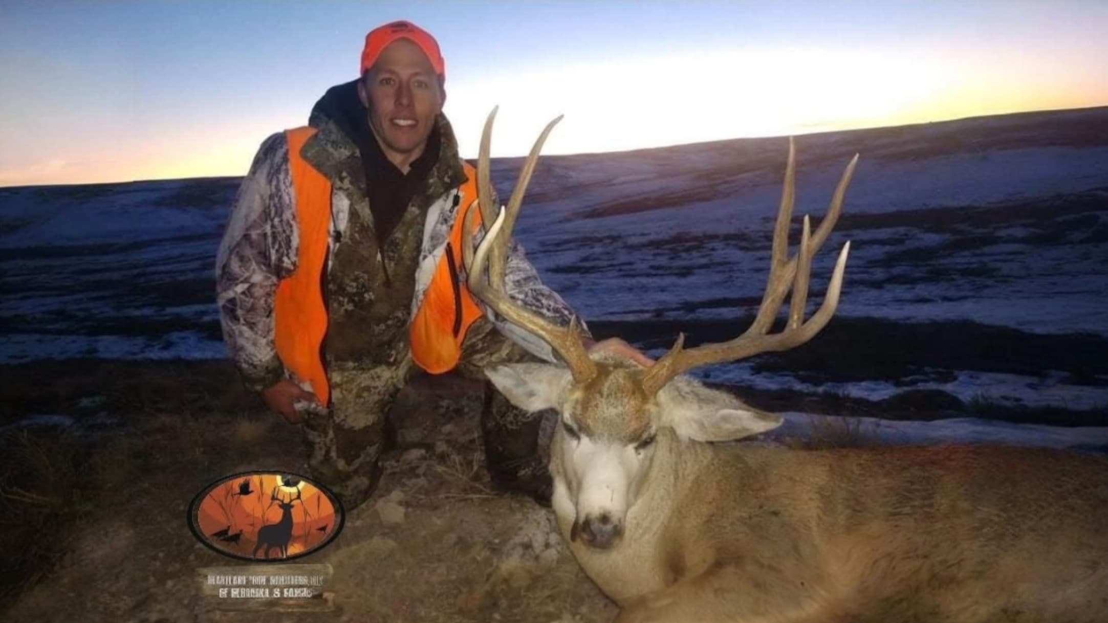 Man in hunting gear kneels beside a large buck with impressive antlers. The deer has light-colored fur. Sunrise or sunset ...