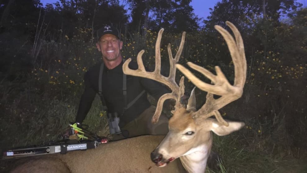Man with a big buck. Man in black clothing, buck with large antlers, bow in front. Dark evening in field with some flowers.