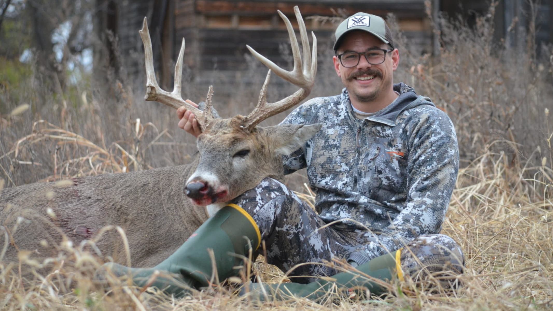 Man with mustache and hat smiles, posing with a large buck. The deer rests on his lap; it has a large rack and blood on it...