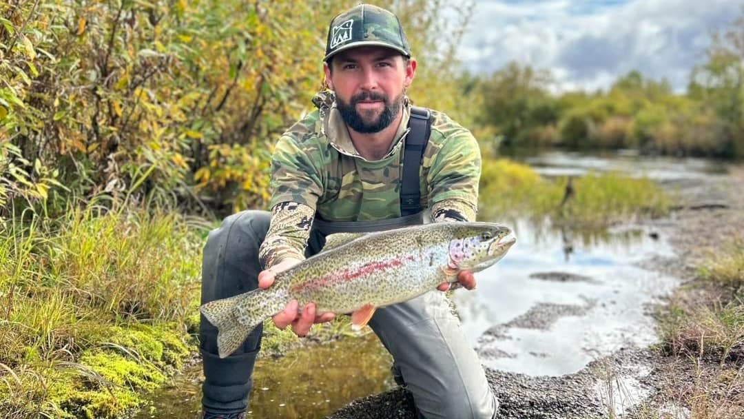 Man kneeling, holding a rainbow trout. He's in camo gear and a cap. Trout has distinctive red stripe. Setting is a marshy ...