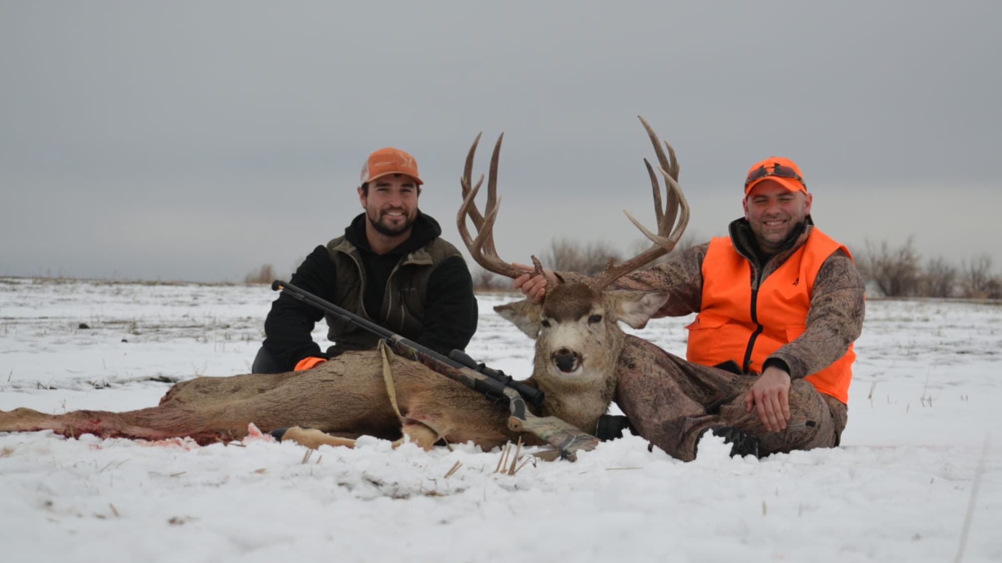 Two hunters pose with a large buck in the snow. Both wear orange hats and vests. The buck has large antlers, rifle alongside.