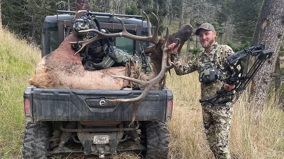 Hunter with large elk antlers in the back of a UTV, holding a compound bow.