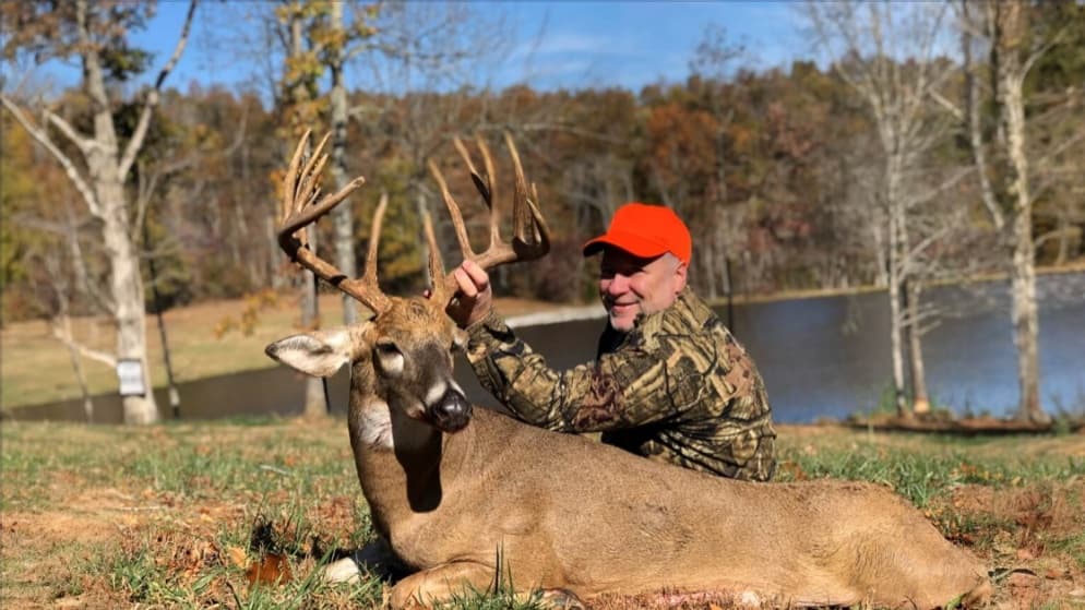 Man in camo and orange hat with a large buck, antlers prominent. The deer lies in grass near trees and a pond in fall.