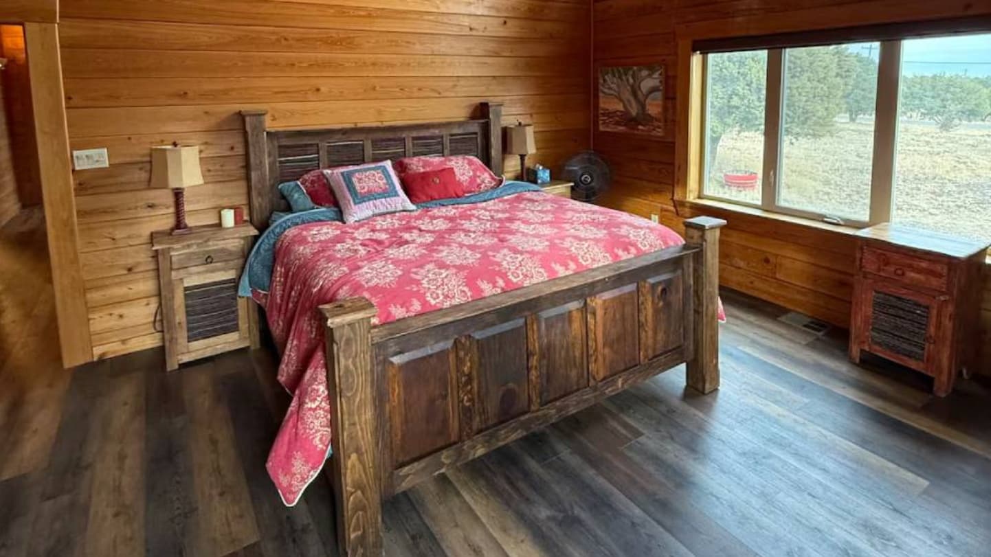 Rustic bedroom featuring a wooden bed with floral bedding. Wood paneling covers walls. Two bedside tables with lamps and a...