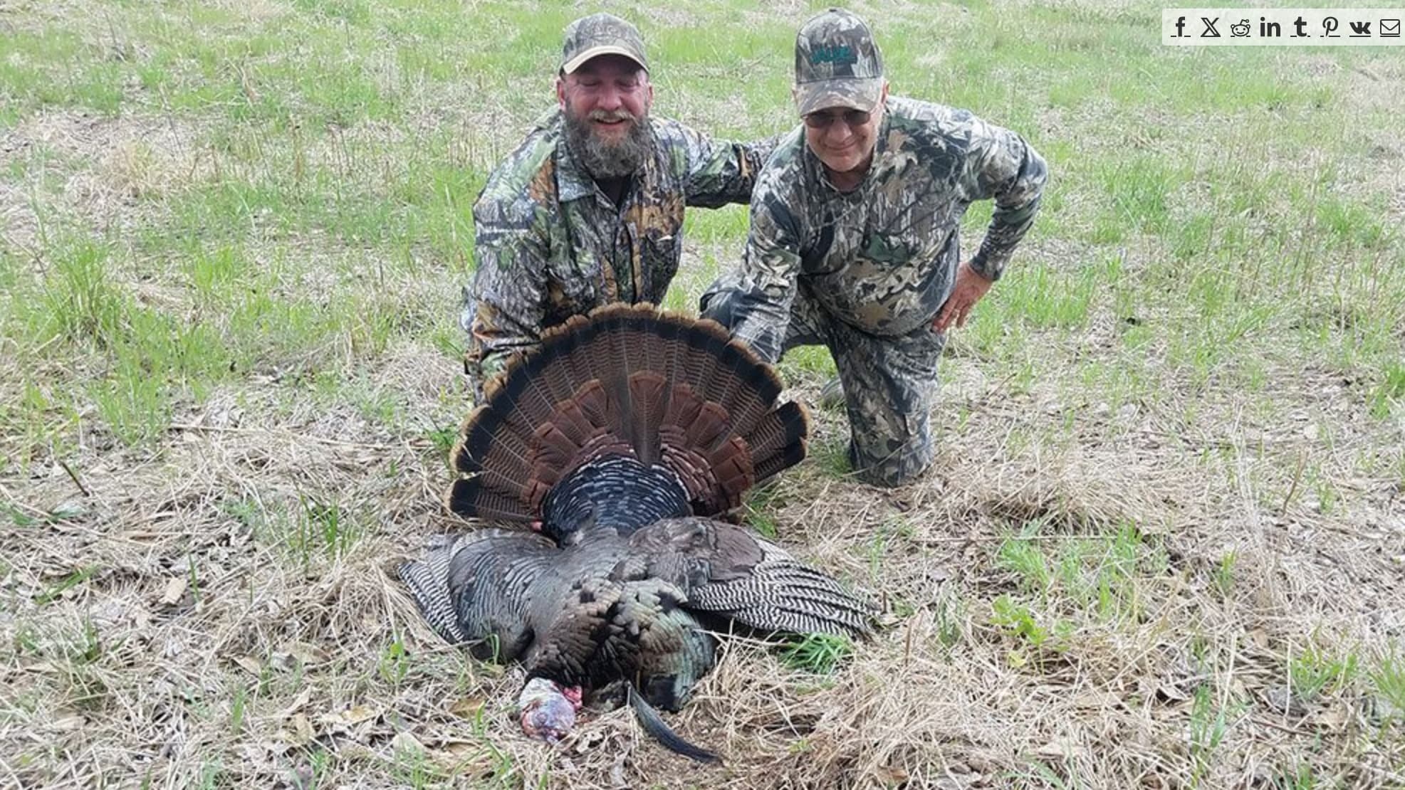 Two hunters in camouflage pose with a large turkey's fanned tail feathers.