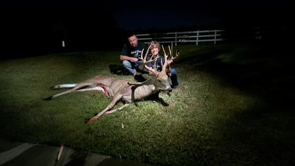 A large buck with large antlers lies on the grass as a man and a boy kneel behind it. The scene is lit with artificial light.