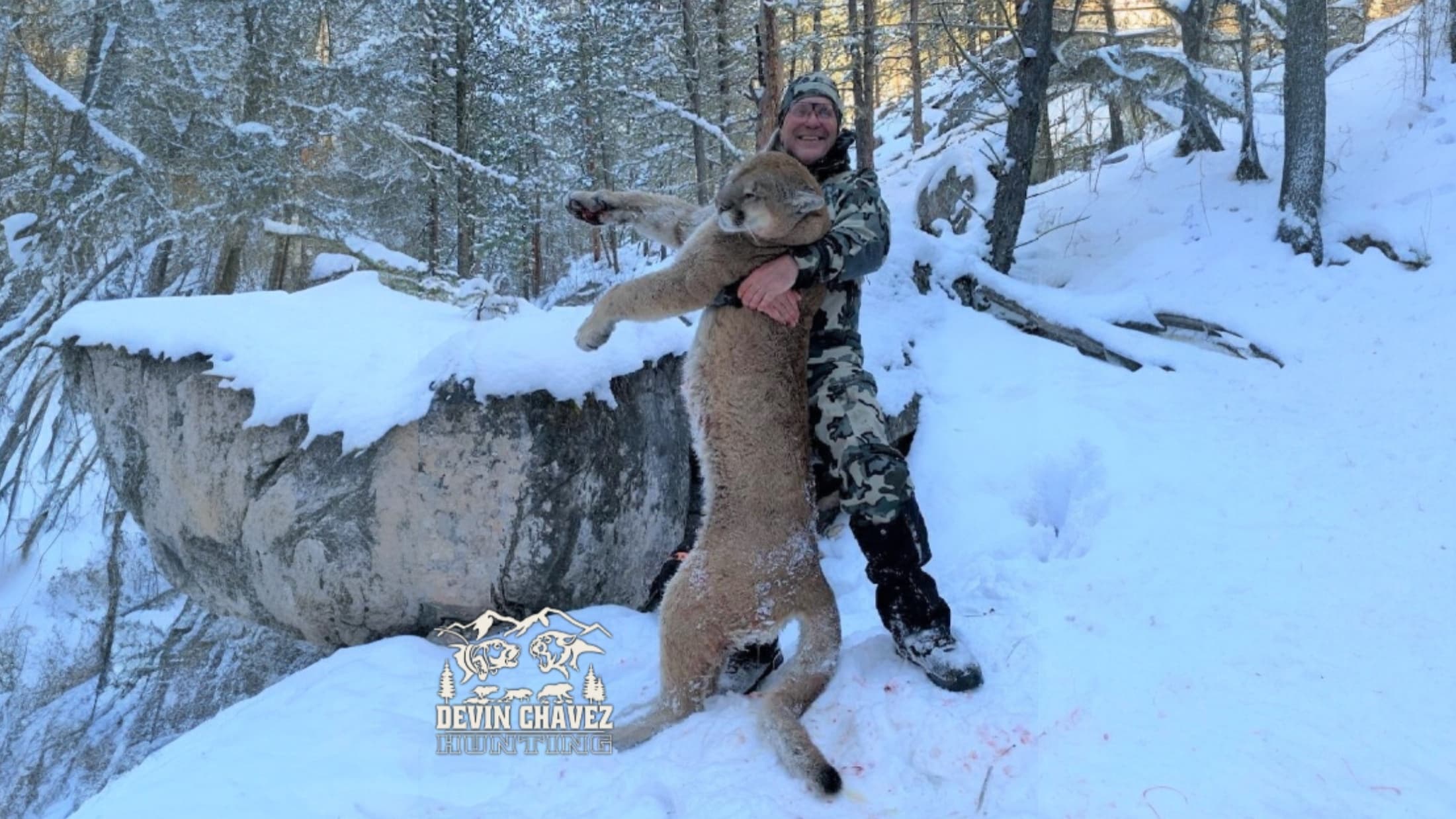 Hunter in camo holds a large dead mountain lion in snowy forest. Logo on snow. Red stains visible.