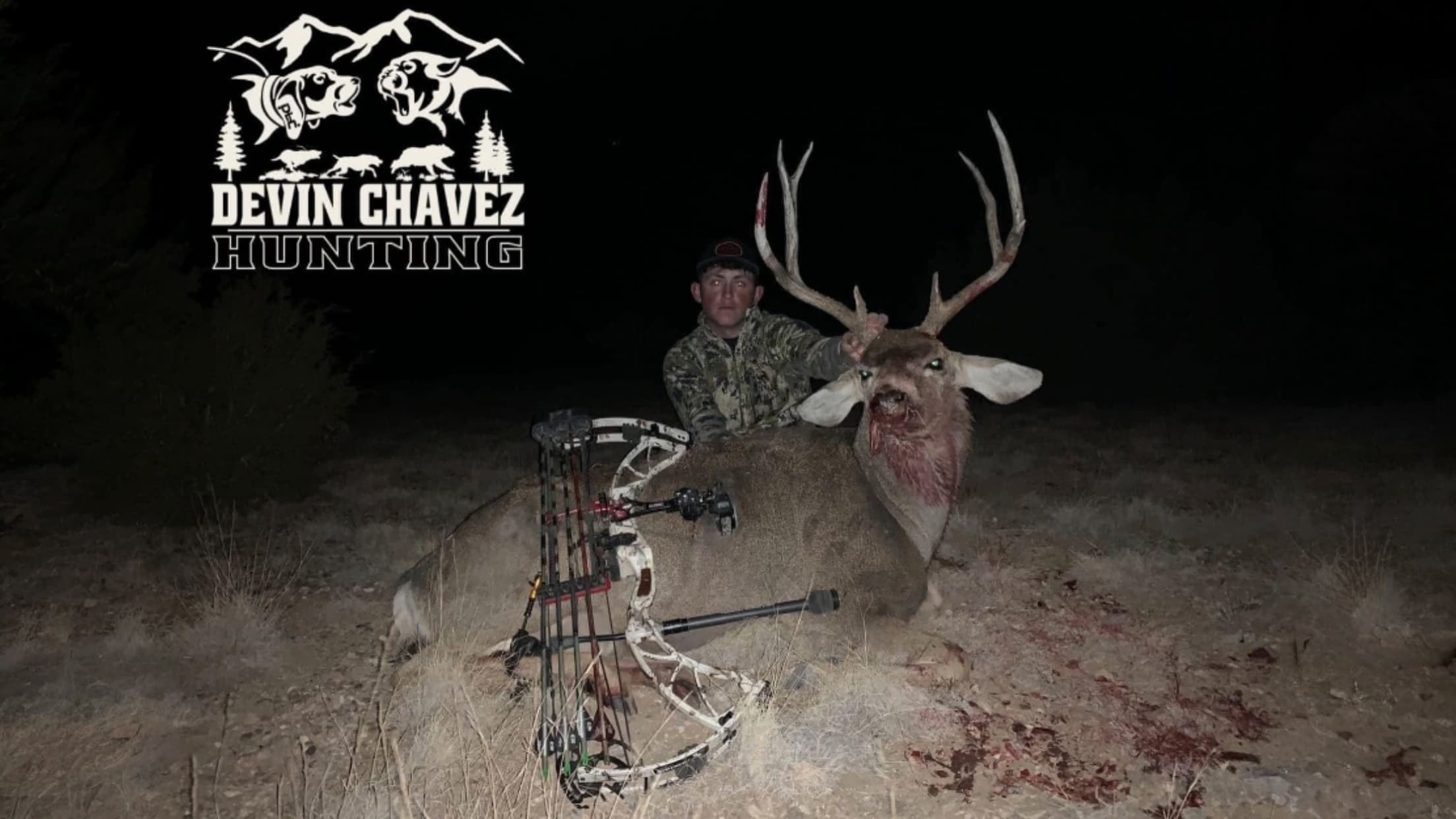 Hunter posing with a harvested deer. The deer has large antlers, blood around its face. A bow is lying next to the deer. T...