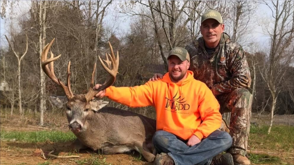 Two men pose with a large buck. One man in orange sweatshirt kneels holding the antlers, the other in camo stands next to ...