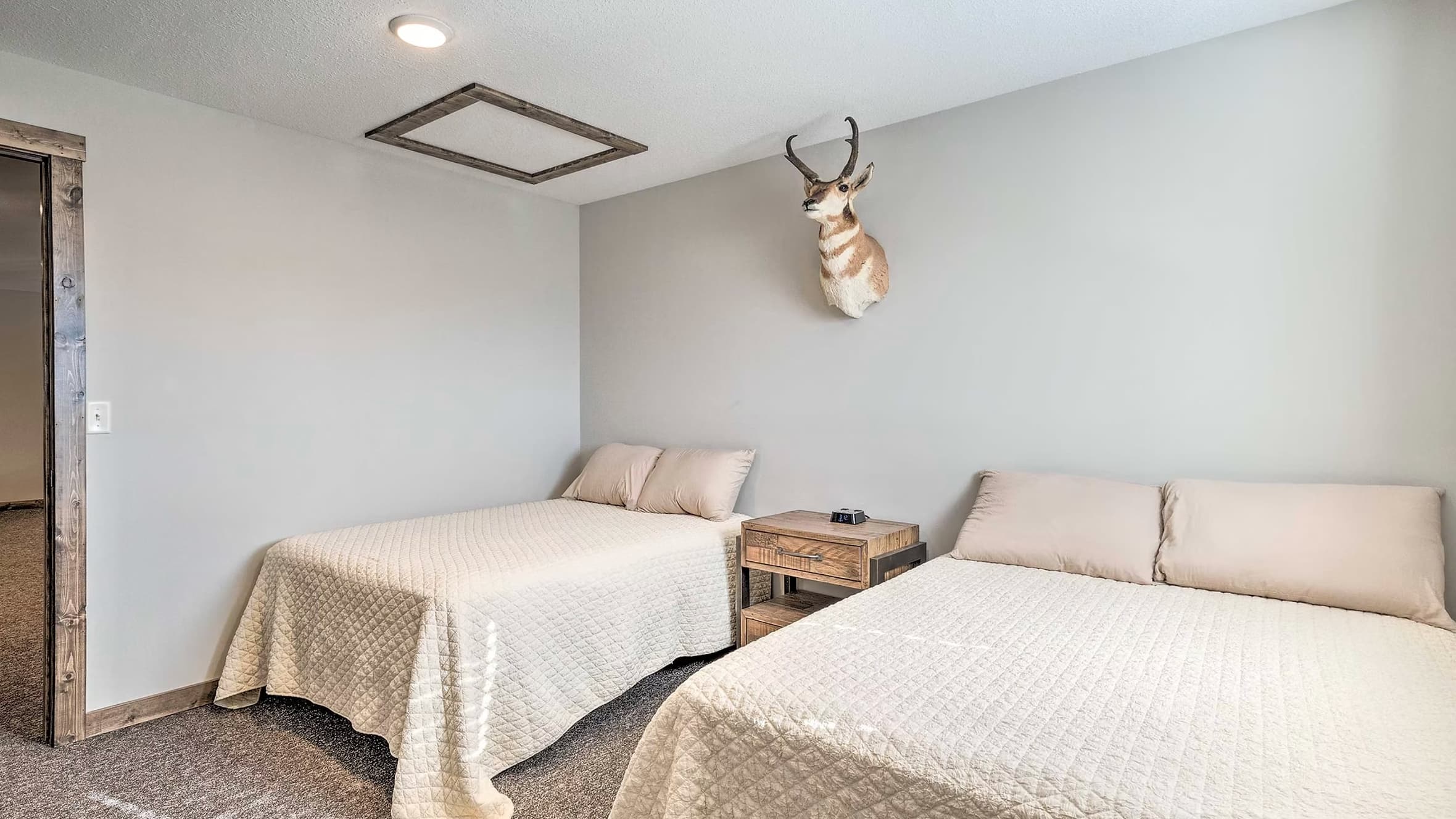 Bedroom with two twin beds, tan bedding, wooden nightstand, and pronghorn taxidermy on the wall. A square ceiling trim is ...