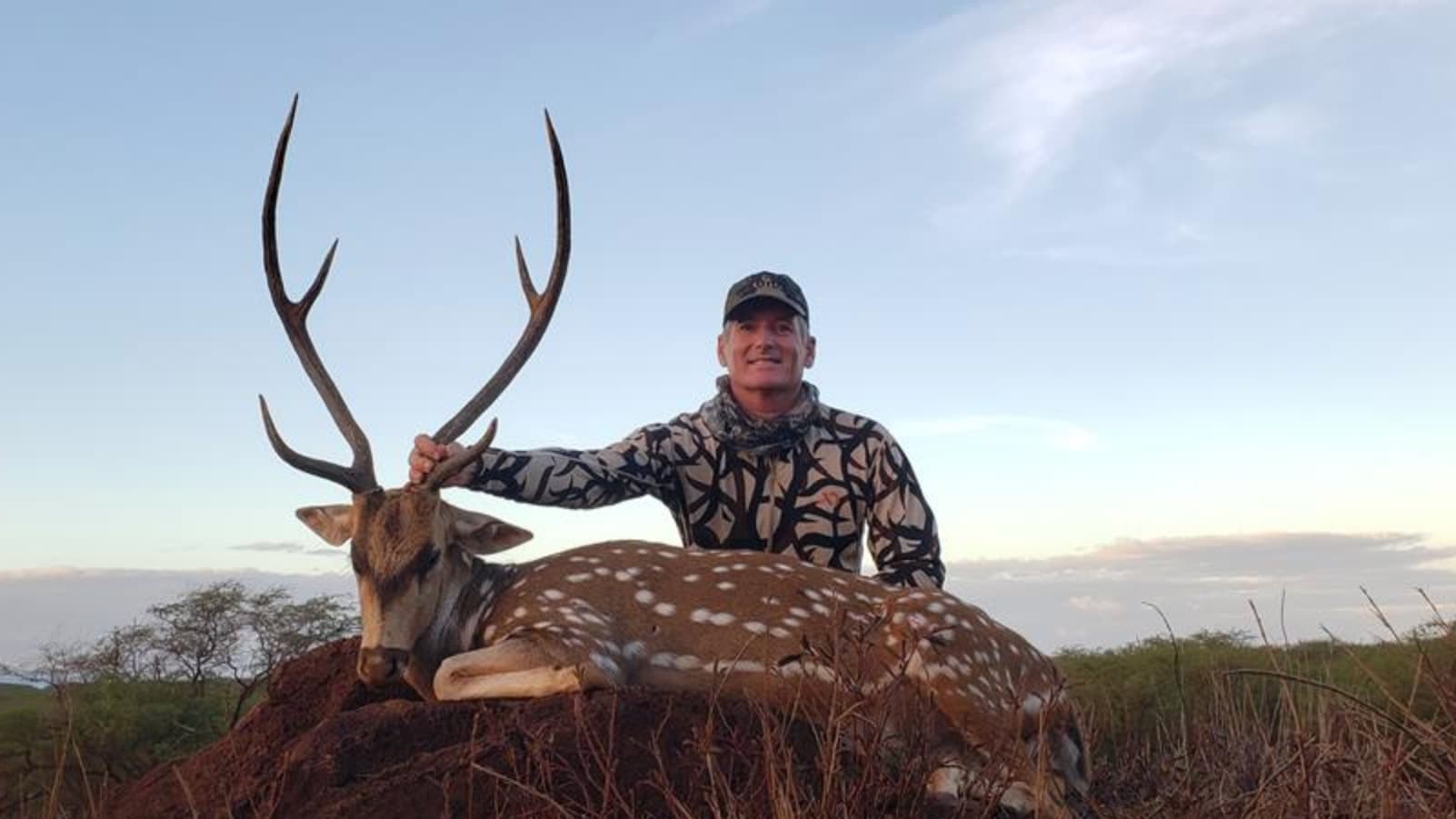 Man in camo holds antlers of a spotted deer lying on the ground. Clear sky in the background, landscape with dry brush and...