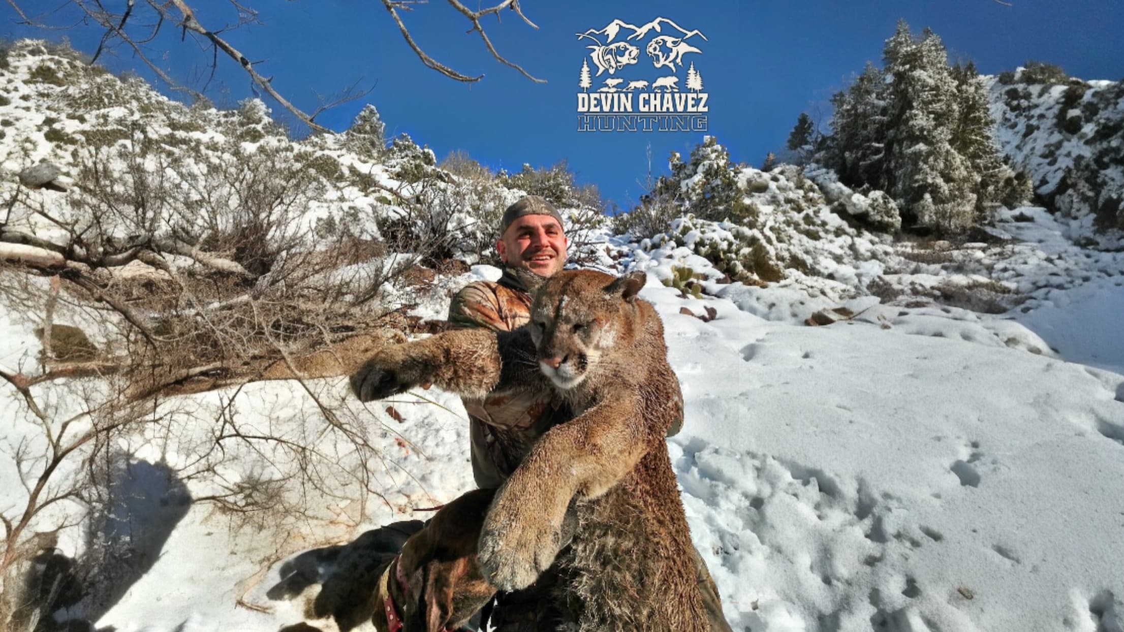 Man holds a large cougar in a snowy, mountainous landscape. The man smiles and the animal is held close, with a bright blu...